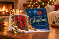 A festive Christmas and New Year greeting card displayed on a wooden table in a cozy living room setting with a fireplace, decorated Christmas tree, and a mug in the background. The card features a dark blue background with gold and white text reading 'Merry Christmas & A Happy New Year', decorated with snowflakes, a red sun, and an illustration of a wrapped gift with a red bow.
